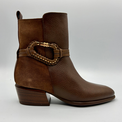 A brown leather ankle boot with a textured pebbled finish, a stacked block heel, and a decorative braided strap with a gold-tone buckle around the ankle.
