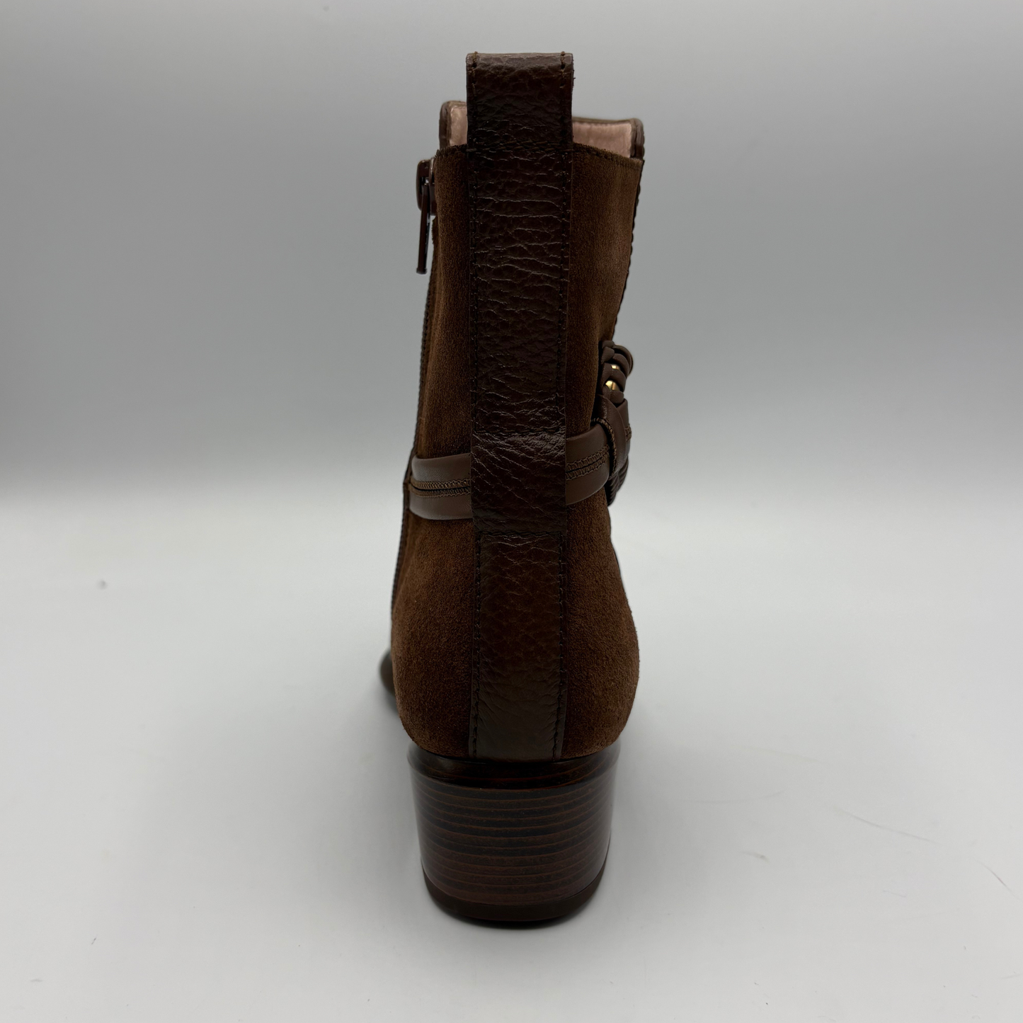 A brown suede ankle boot shown from the back, featuring a textured heel, a vertical pull tab, and a side buckle strap, photographed on a neutral gray background.