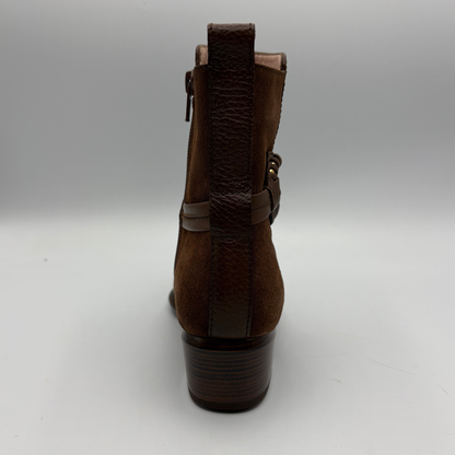 A brown suede ankle boot shown from the back, featuring a textured heel, a vertical pull tab, and a side buckle strap, photographed on a neutral gray background.