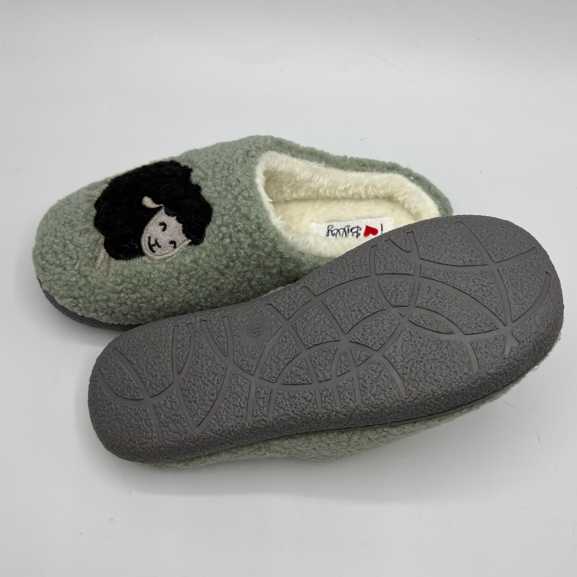 A pair of fuzzy slip-on slippers on a light gray background. One slipper is shown from the top, featuring a sage green exterior with a black sheep character embroidered on the side and a cream sherpa lining.
