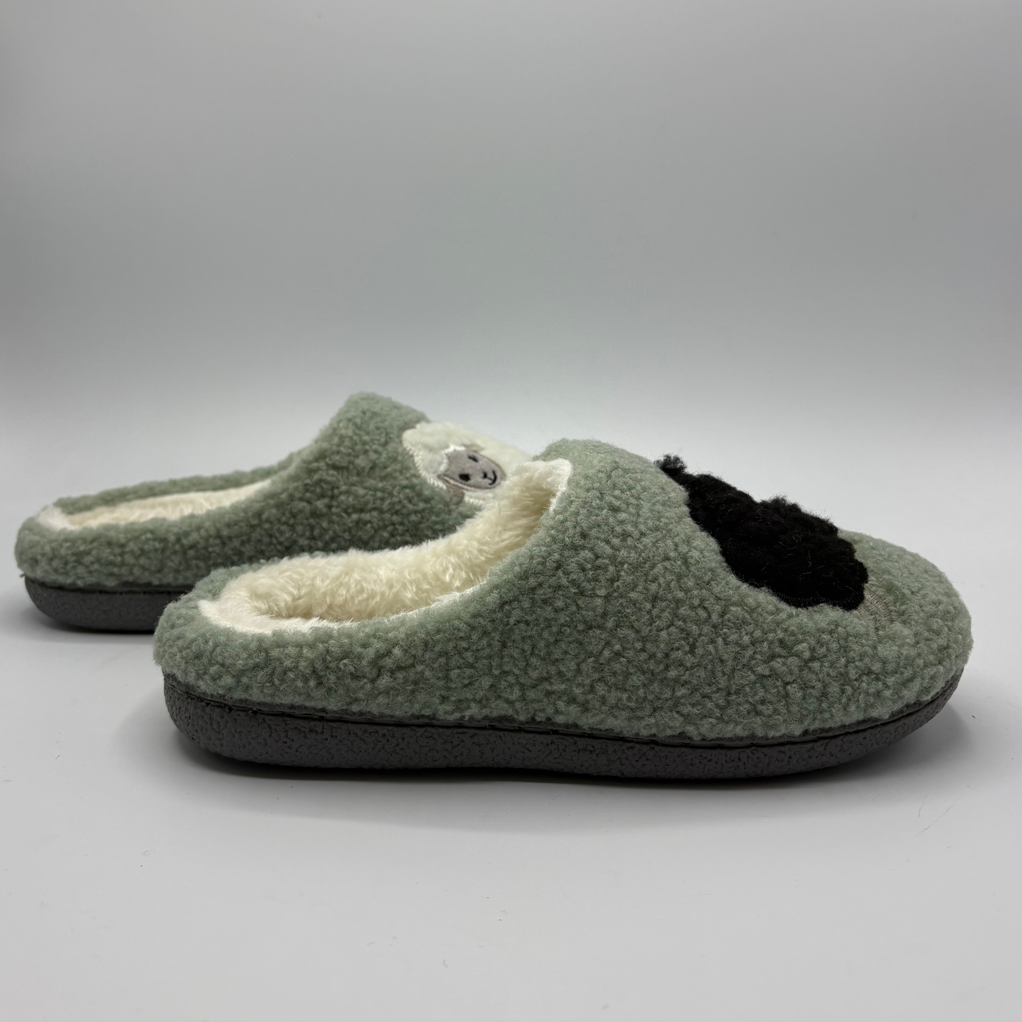 A pair of plush fleece slip-on slippers in sage green, featuring a cream sherpa lining, with a black sheep detail on the toebox