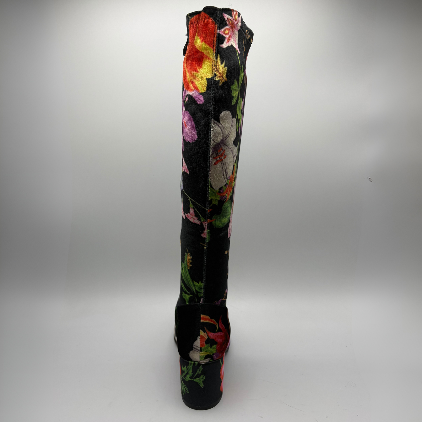 A single knee-high boot with a black base and a vibrant, multicolored floral print, shown from the back against a neutral gray studio background.