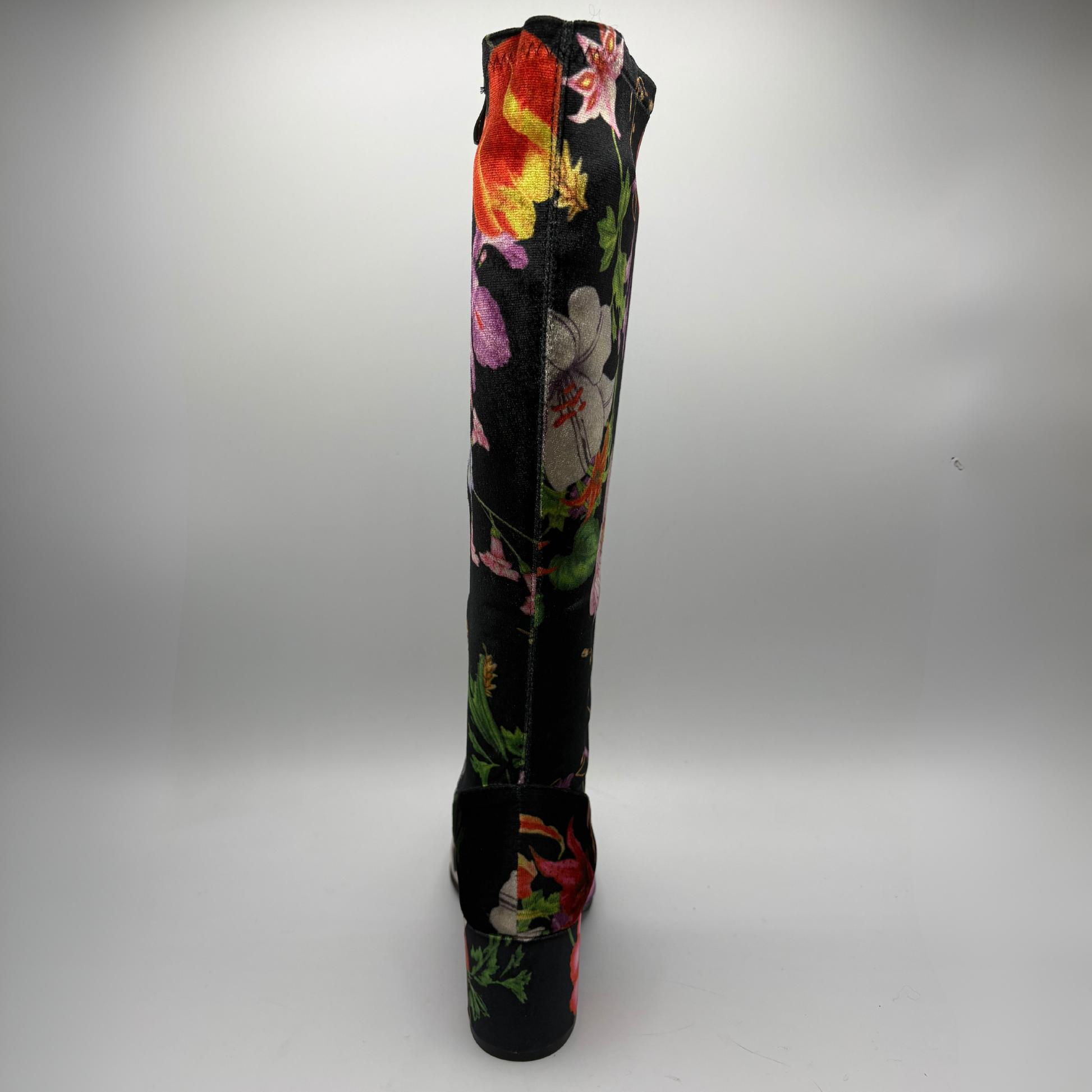 A single knee-high boot with a black base and a vibrant, multicolored floral print, shown from the back against a neutral gray studio background.