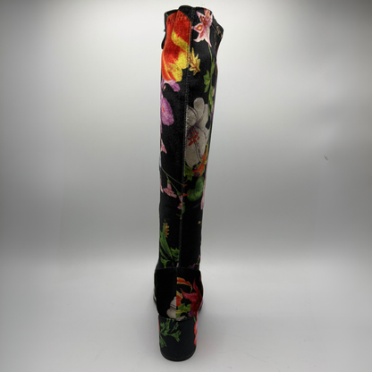 A single knee-high boot with a black base and a vibrant, multicolored floral print, shown from the back against a neutral gray studio background.