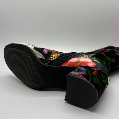 A close-up studio shot of a black velvet boot featuring a vibrant multicolor floral print and a chunky black block heel, with the sole shown in the foreground.