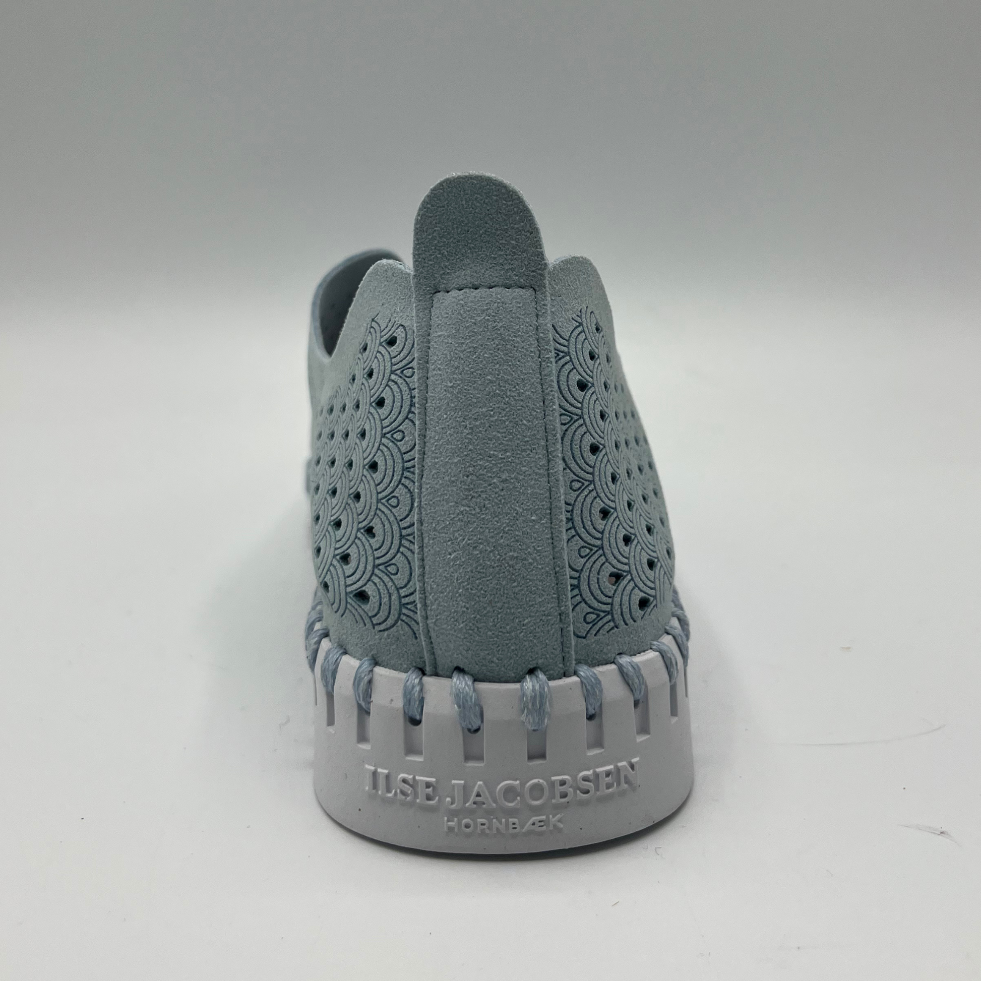 Back facing view of ash blue slip on shoe with an intricate laser cut upper and white rubber outsole.