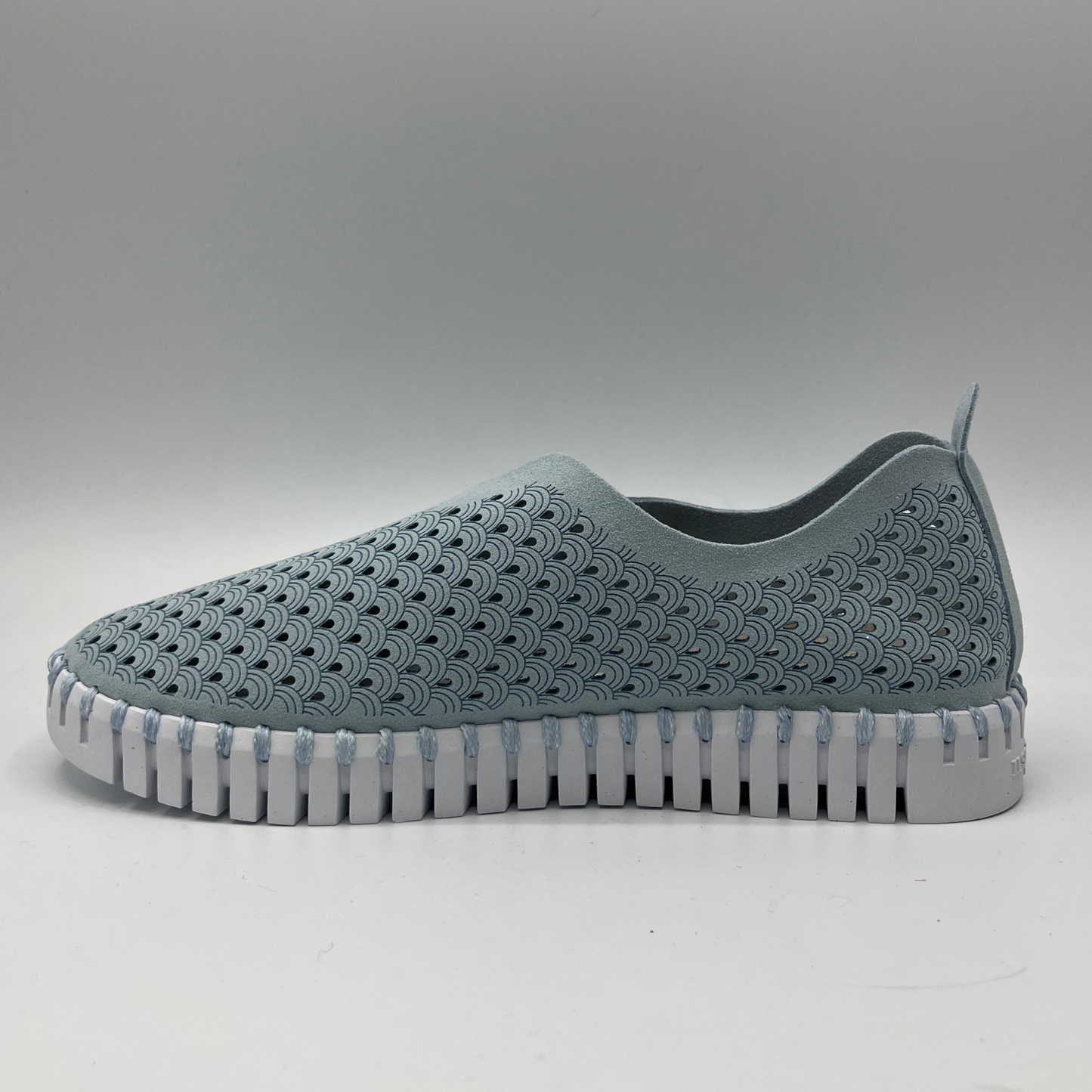 Left facing view of ash blue slip on shoe with an intricate laser cut upper and white rubber outsole.