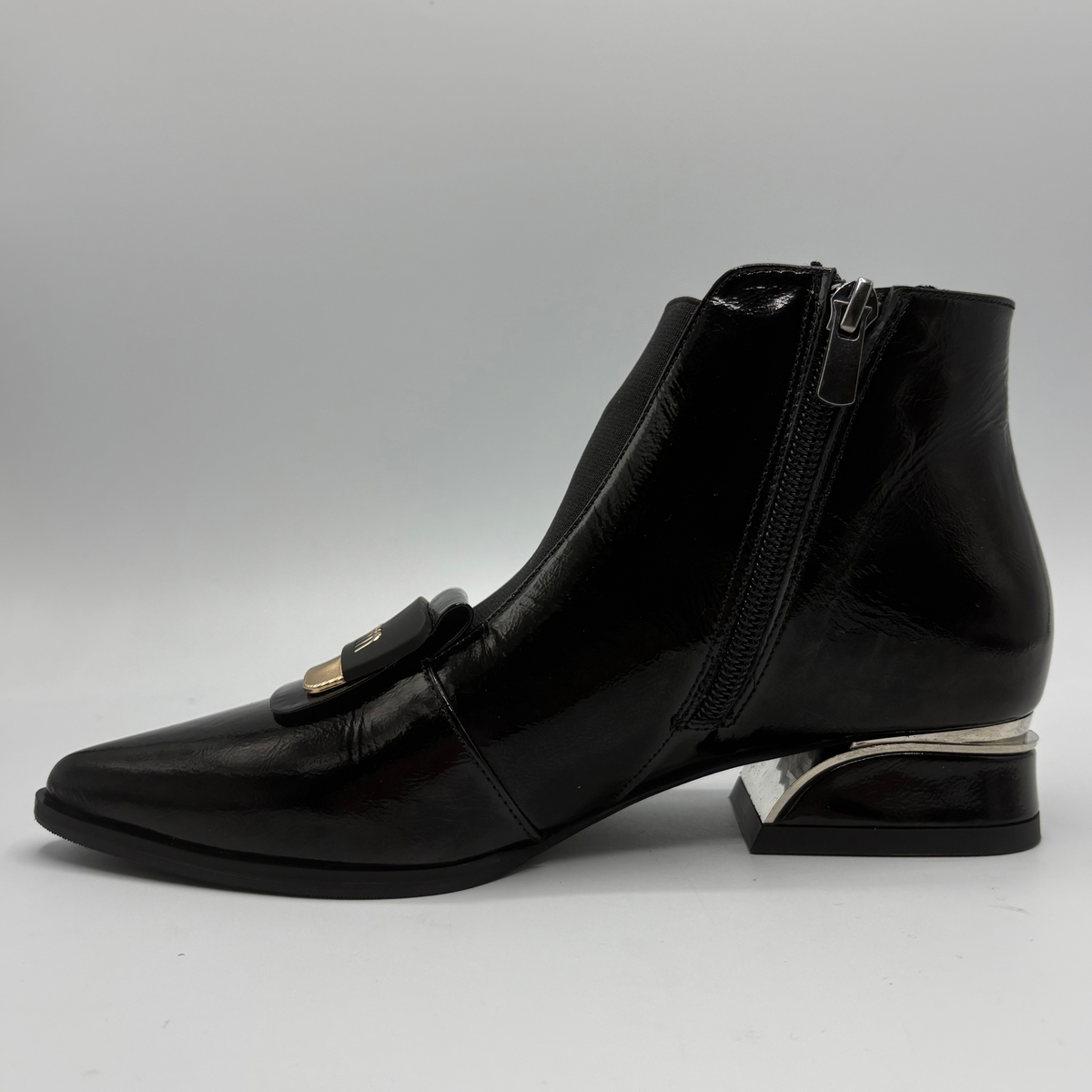 A black patent leather ankle boot with a pointed toe, side zipper, and a decorative gold-tone buckle detail on the vamp, shown against a neutral gray background.
