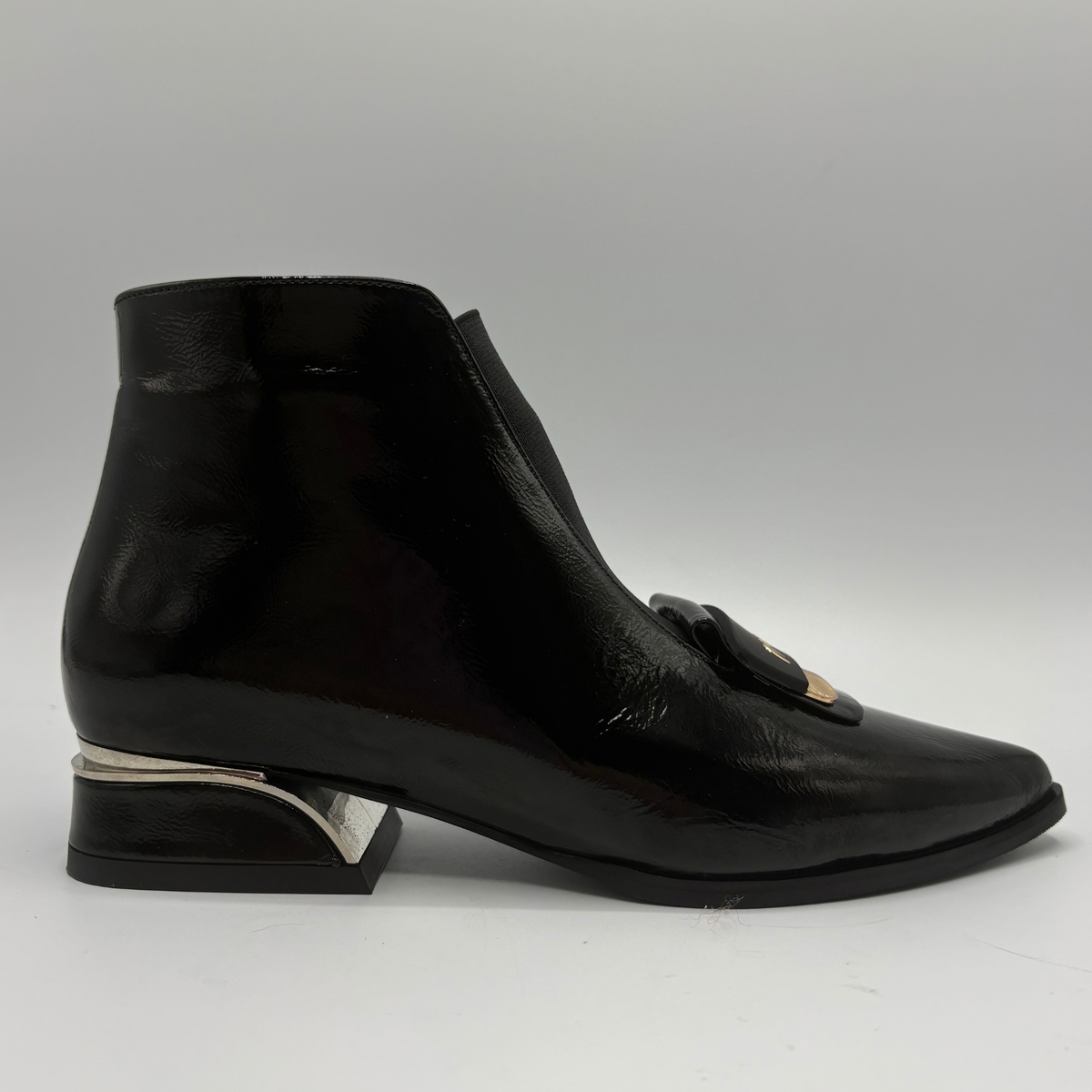 A single black leather ankle boot with a glossy finish, a low block heel with a silver metallic accent, and a decorative black bow with a gold metal center on the toe, shown against a neutral gray background.