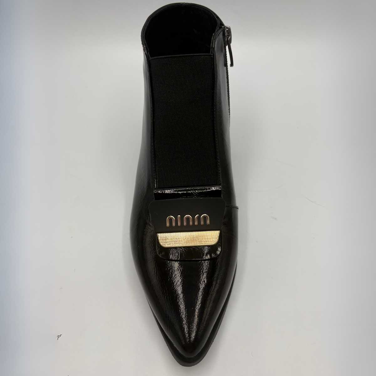 A single black pointed-toe ankle boot with a side zipper and a gold-tone logo plaque on the front, photographed from a top-down angle on a light gray background.