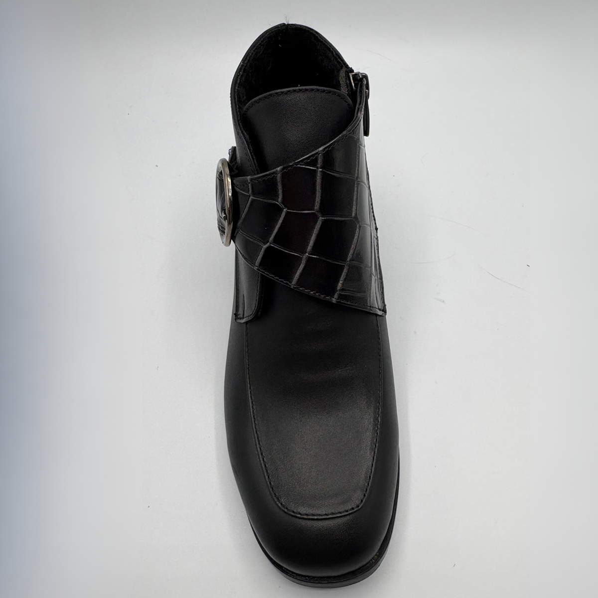 Top view of a black leather ankle boot with a croc textured strap and a circular buckle