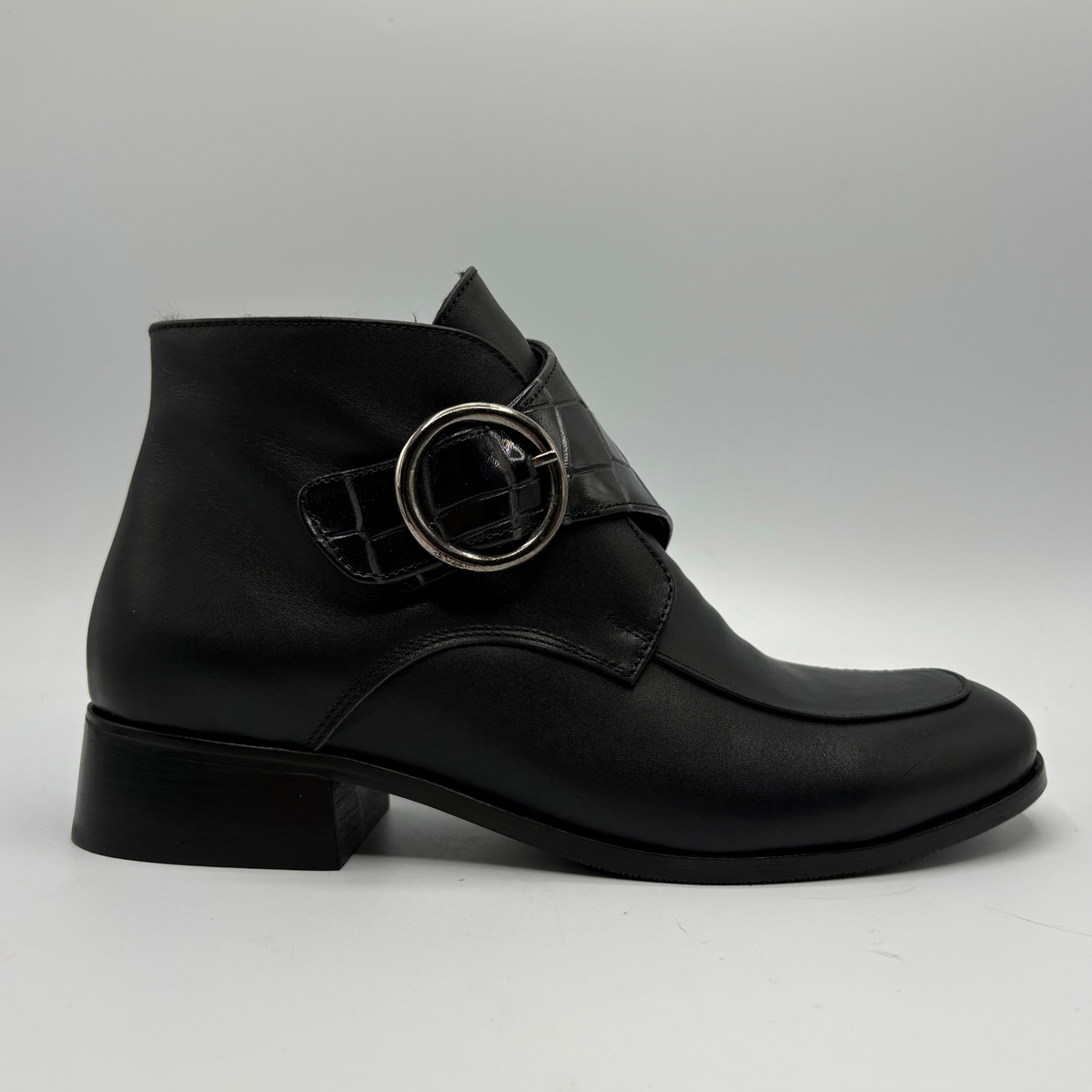 Right facing view of a black leather ankle boot with a croc textured strap and a circular buckle