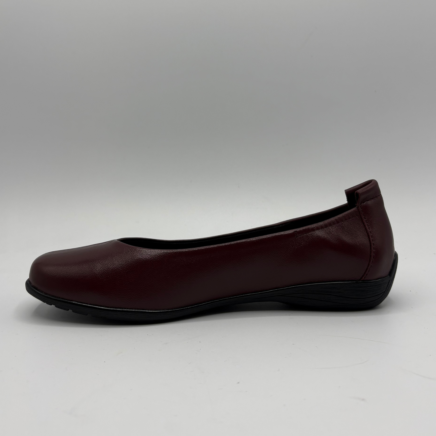 A single women's flat shoe in burgundy leather with a black sole, shown in a side profile view against a neutral gray background.