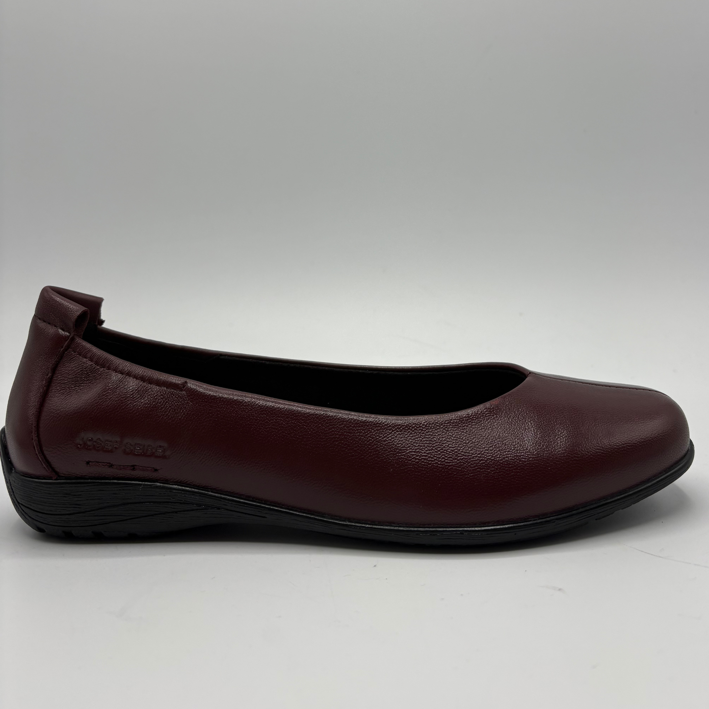 A single women's flat shoe in burgundy leather with a black sole, shown from a side profile view against a light gray background.