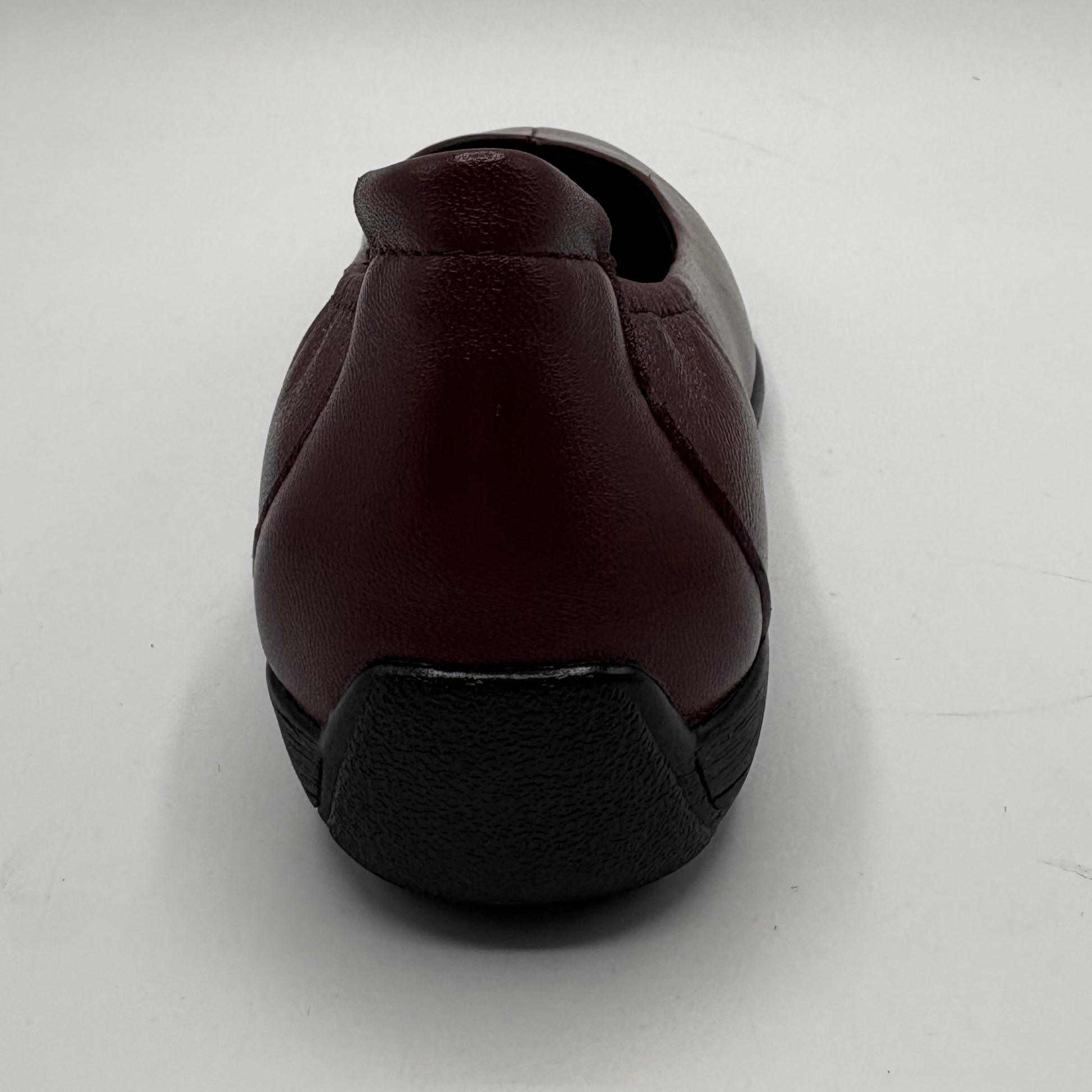 A single slip-on shoe shown from the back, featuring a dark burgundy leather upper and a black textured sole, set against a plain light gray background.