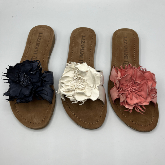 View of three slip on sandals each with a handmade, hand dyed leather flower. The left one is black, the middle is off white and right right is a coral pink.