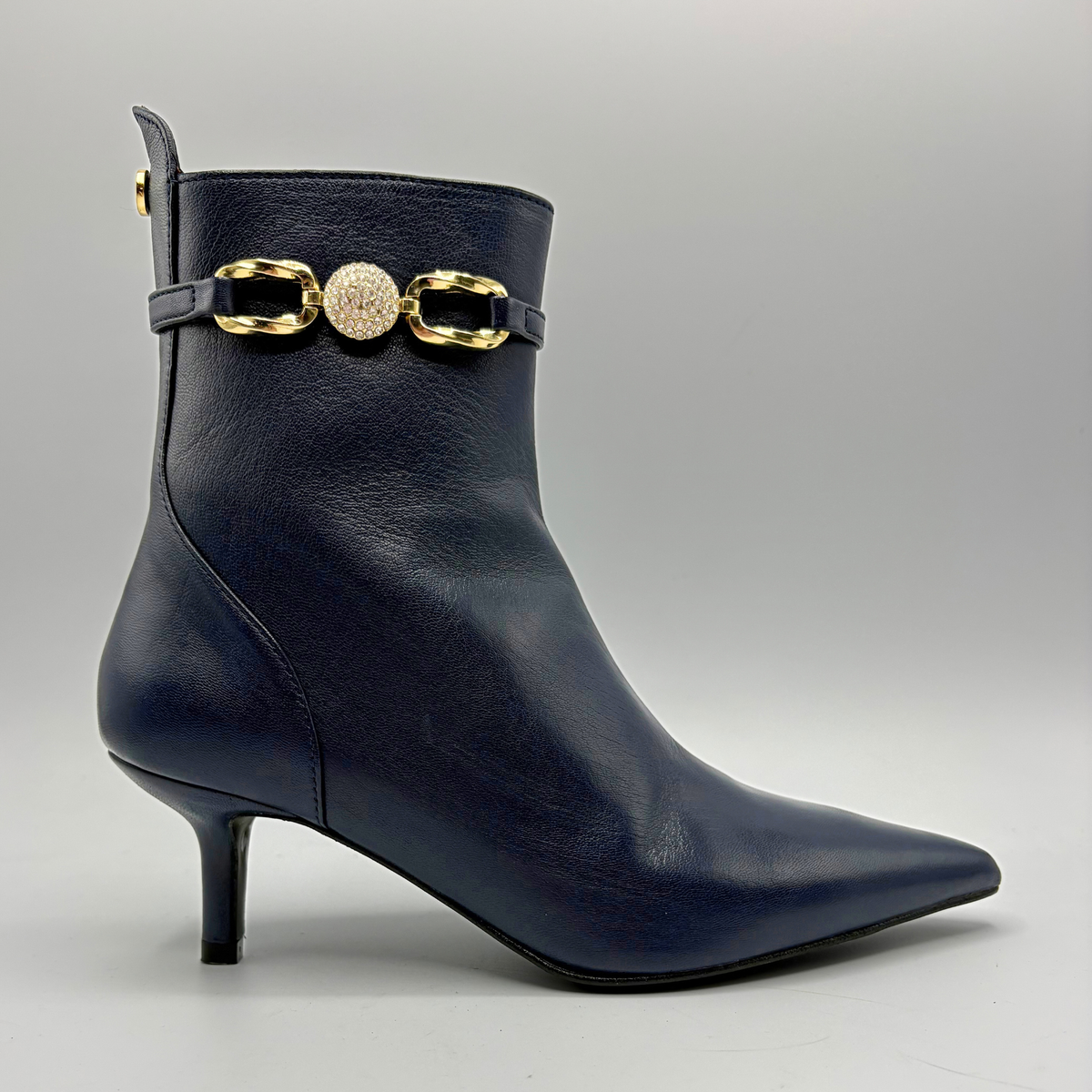 A navy blue leather ankle boot with a pointed toe, kitten heel, and a decorative gold-tone chain strap with a central textured round charm around the collar, shown against a neutral gray background.