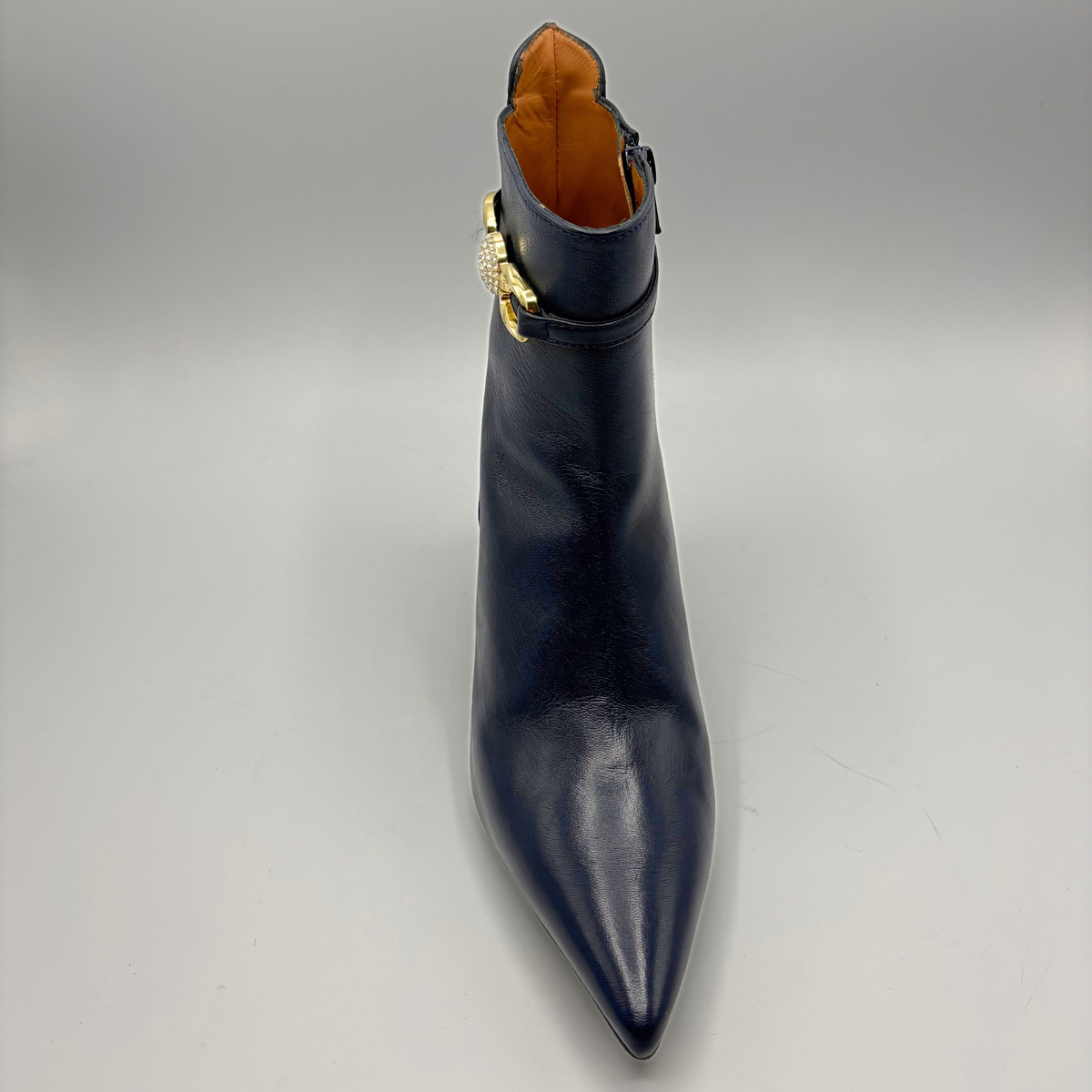 A dark navy blue leather pointed-toe ankle boot with a gold-tone decorative buckle detail on the side, shown against a neutral gray background.