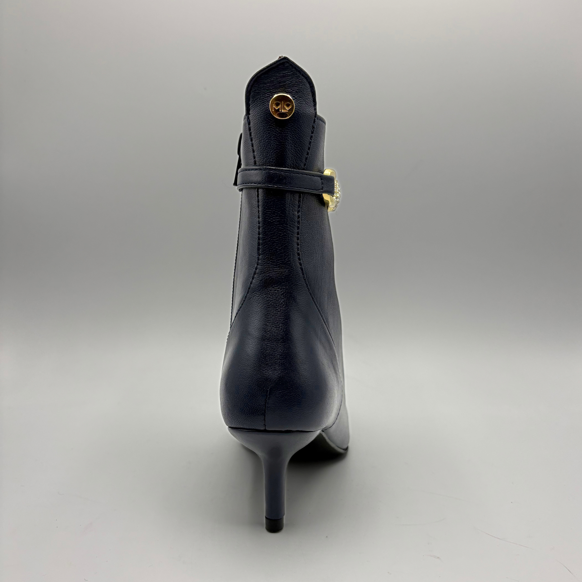 A single navy blue leather ankle boot with a stiletto heel, shown from the back. It features a decorative strap across the back with a gold-tone buckle and a small gold circular emblem near the top collar.