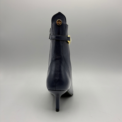 A single navy blue leather ankle boot with a stiletto heel, shown from the back. It features a decorative strap across the back with a gold-tone buckle and a small gold circular emblem near the top collar.