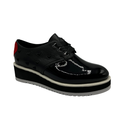 45 degree angled view of black patent leather shoe with red heart on the heel, ribbon laces and thick rubber outsole.