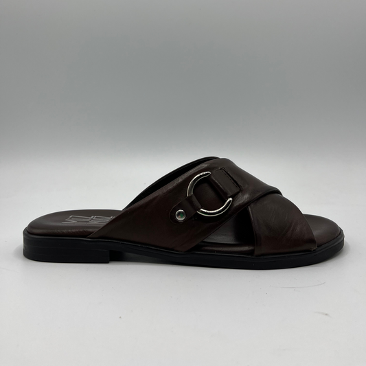 Right facing view of a brown leather slip on sandal with a cross strap design and low heeled outsole. Featuring a silver metal accent on the strap and a cushioned leather insole.