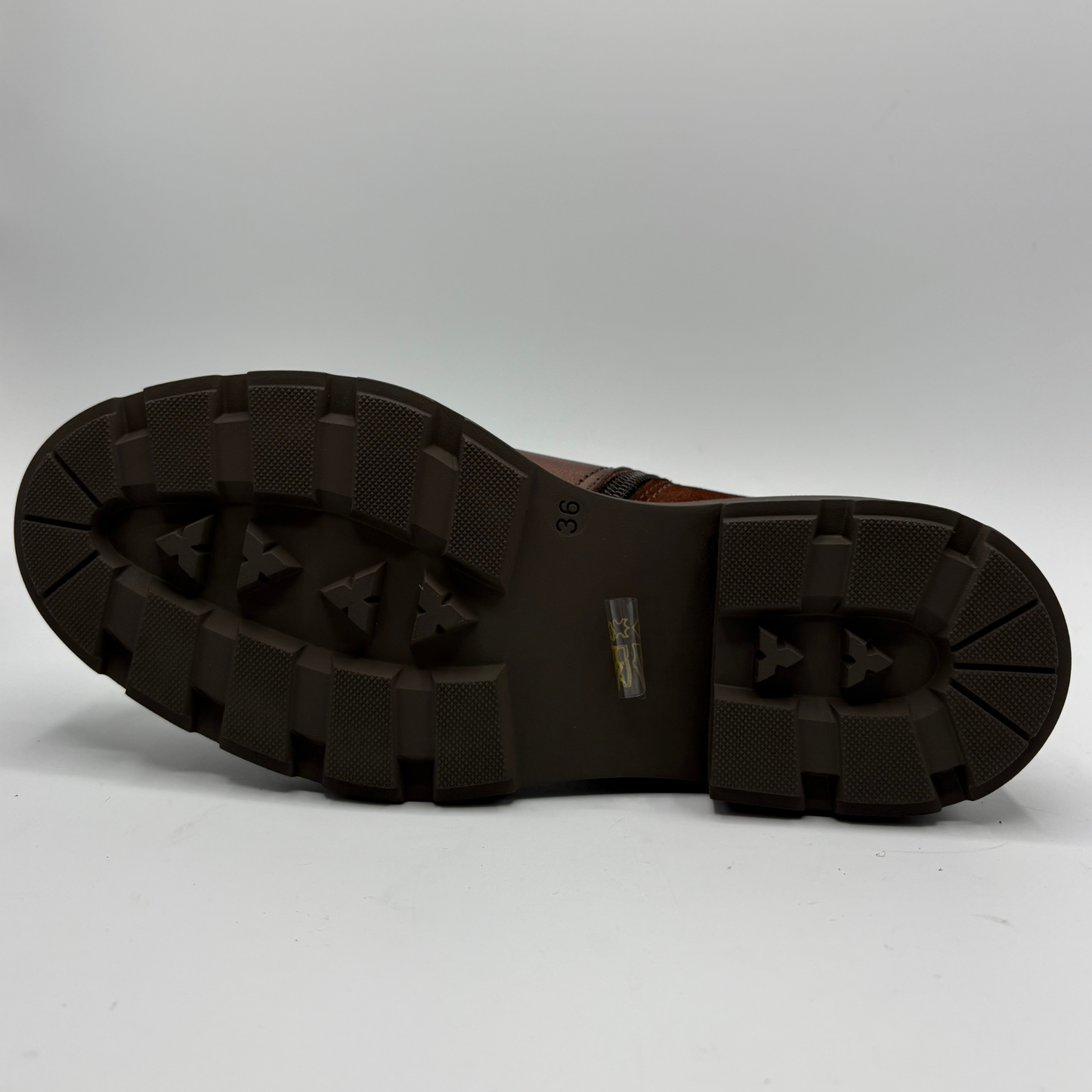 The outsole of a brown leather boot, showing deep tread patterns photographed on a light gray background.