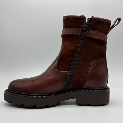 A brick red leather and suede ankle boot with a side zipper, elastic side strap, and a chunky black sole, shown from a side profile view against a light gray background.
