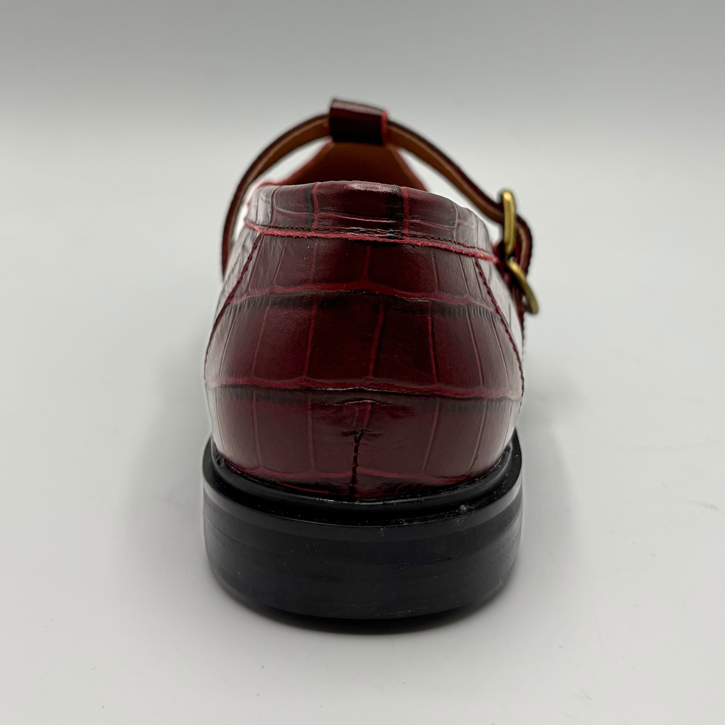 A single red crocodile-embossed leather shoe shown from a rear three-quarter view, featuring a black sole and a gold-tone buckle on a light gray background.