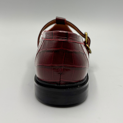 A single red crocodile-embossed leather shoe shown from a rear three-quarter view, featuring a black sole and a gold-tone buckle on a light gray background.