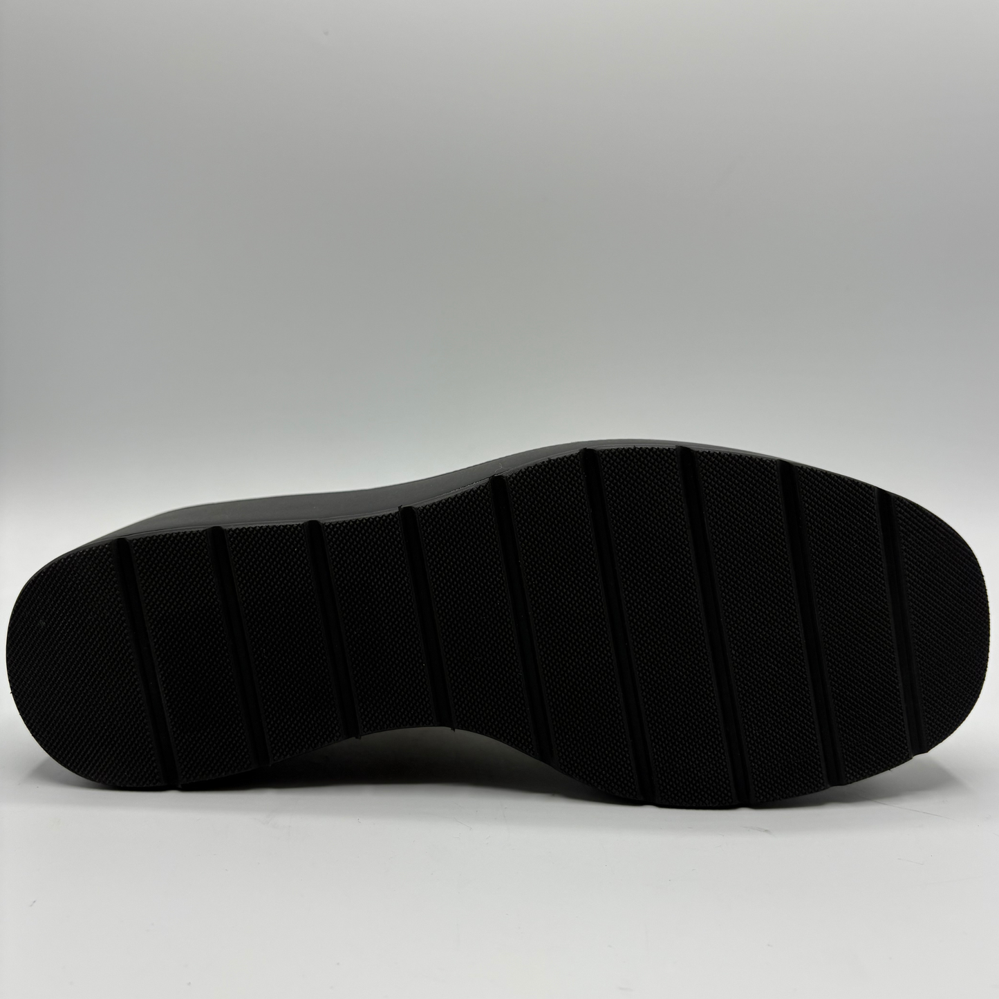 The black textured rubber outsole of a shoe, shown from a top-down view against a light gray studio background.