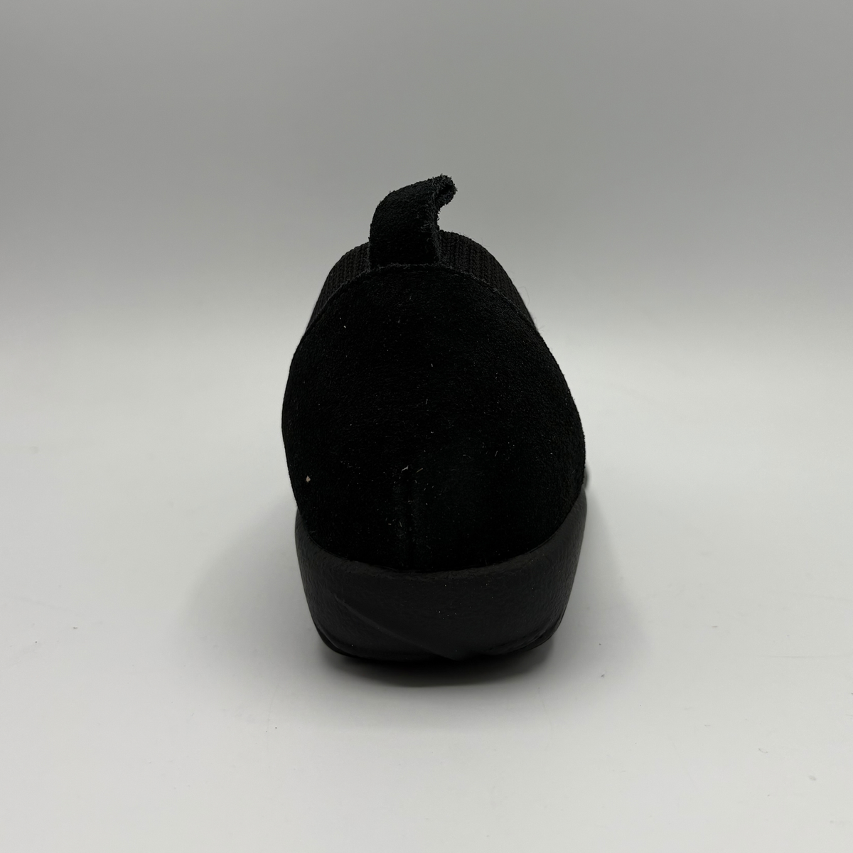 A single black slip-on shoe shown from the back, featuring a textured fabric upper and a thick black sole, set against a neutral gray studio background.