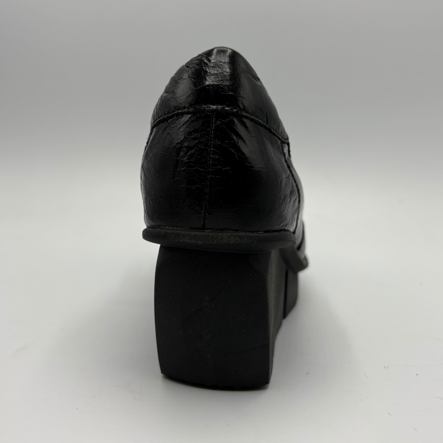 A black leather platform shoe shown from a rear three-quarter angle, highlighting the textured leather upper and chunky platform heel, set against a plain light gray background.