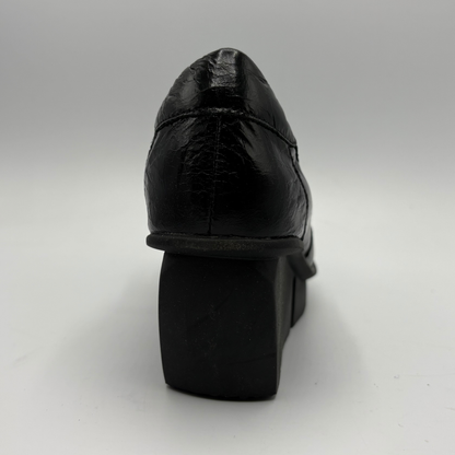 A black leather platform shoe shown from a rear three-quarter angle, highlighting the textured leather upper and chunky platform heel, set against a plain light gray background.