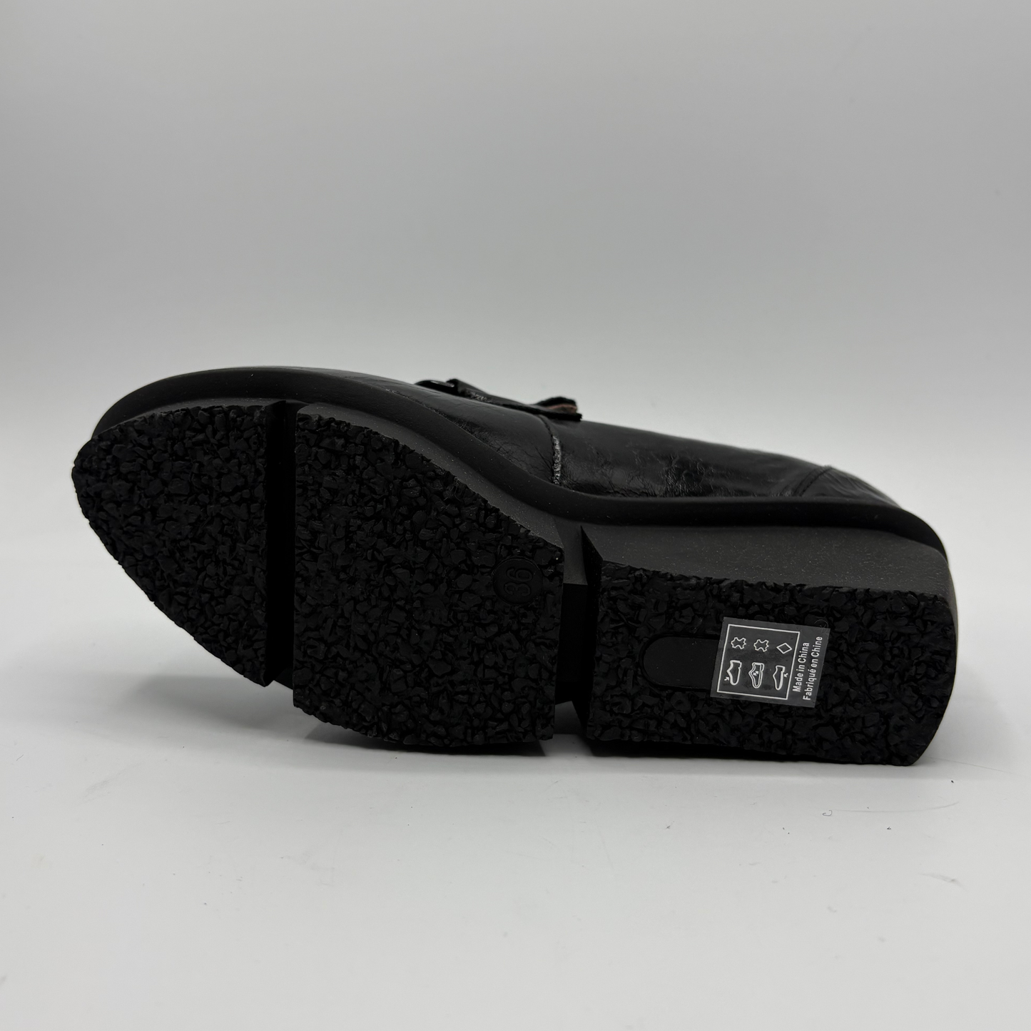 A black leather shoe shown from the sole side, featuring a textured black rubber outsole and a small white brand label on the heel.