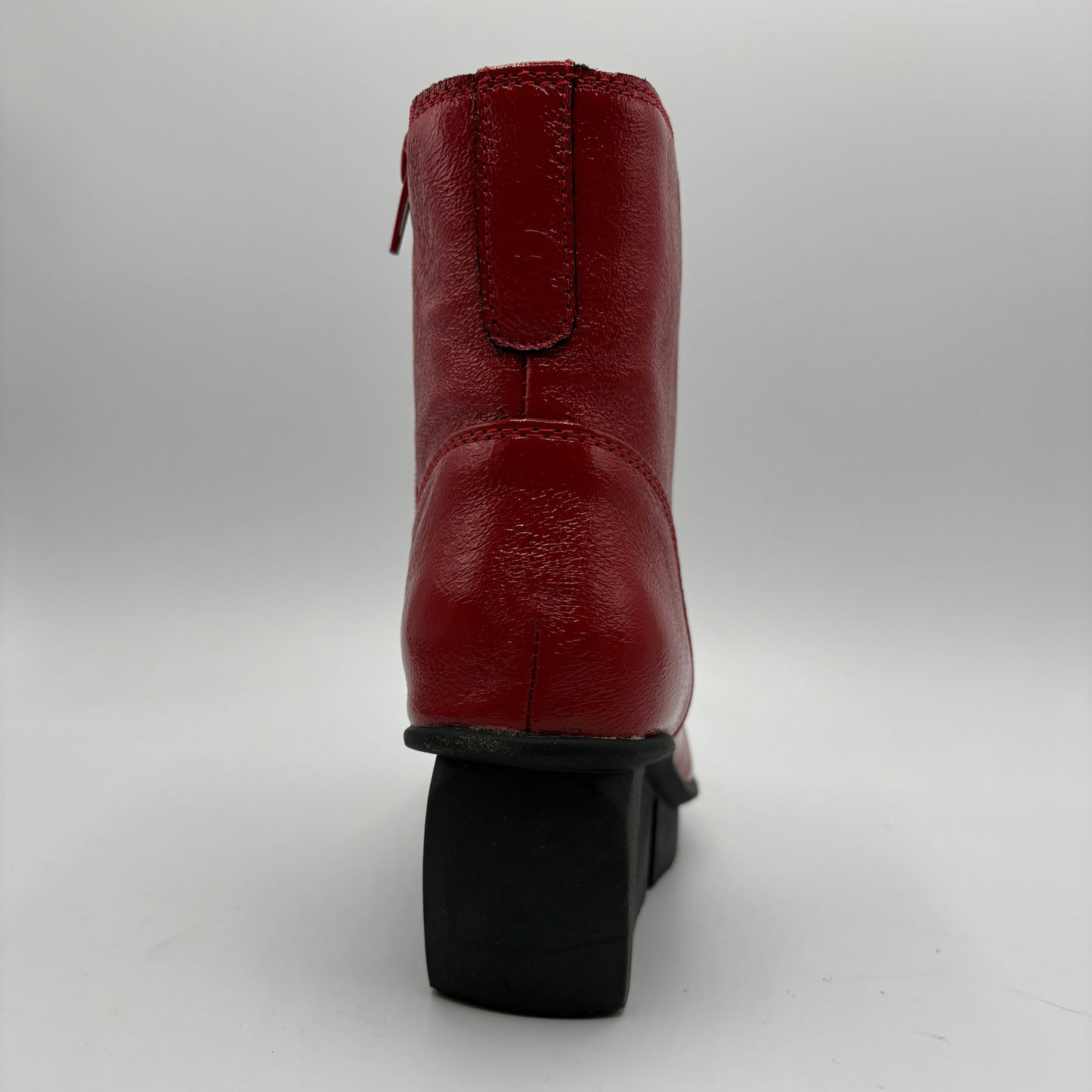 A red leather ankle boot shown from the back, featuring a black block heel and a side zipper, photographed against a neutral gray background.
