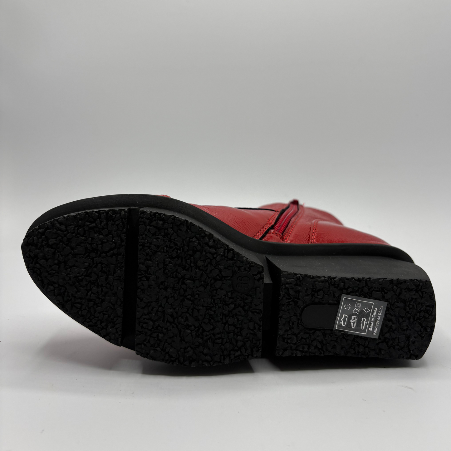 A close-up shot of the sole of a red leather boot, showing the textured black rubber outsole and a small label on the heel, set against a plain light gray background.