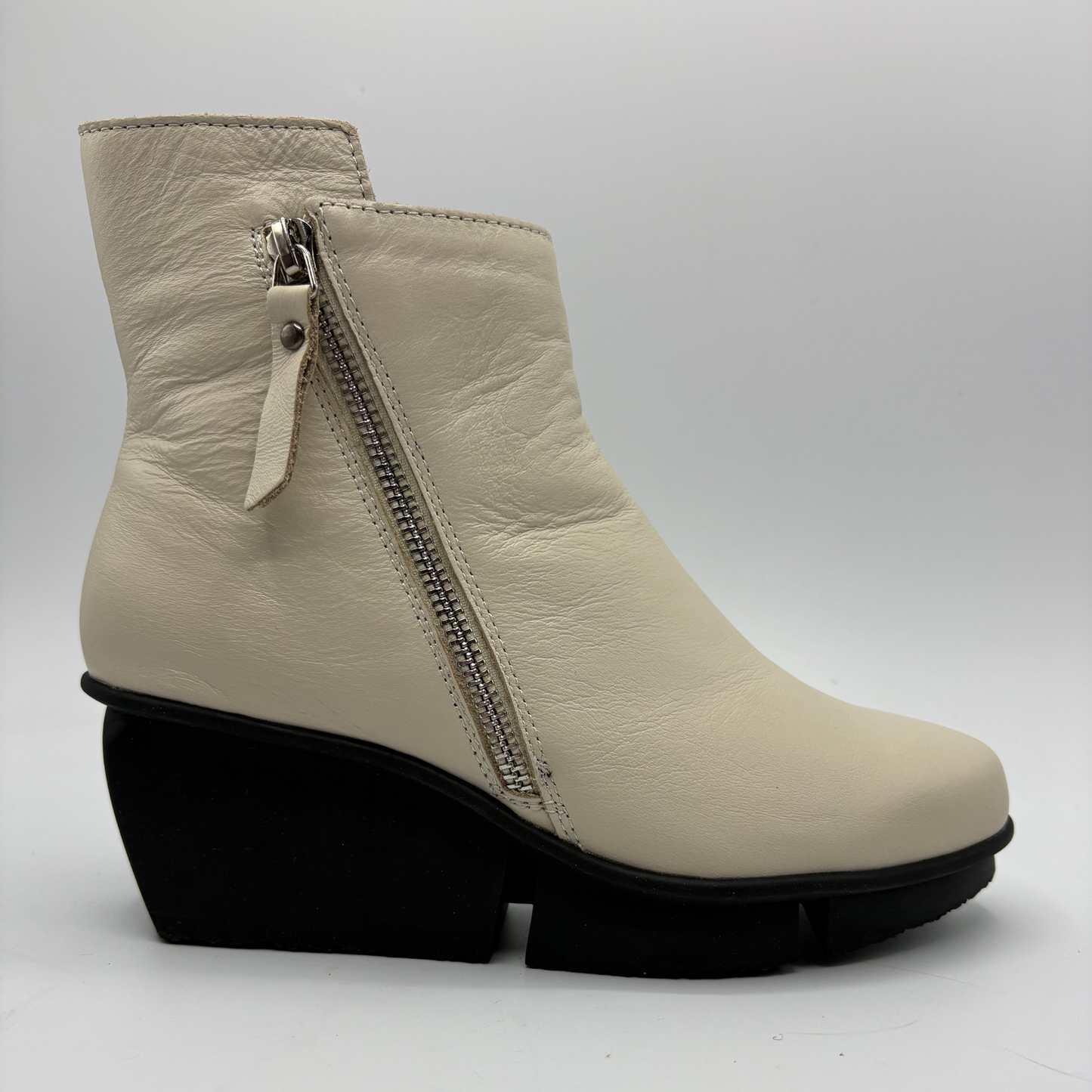 A cream-colored leather ankle boot with a black wedge heel and a silver side zipper, shown from a side profile against a light gray background.