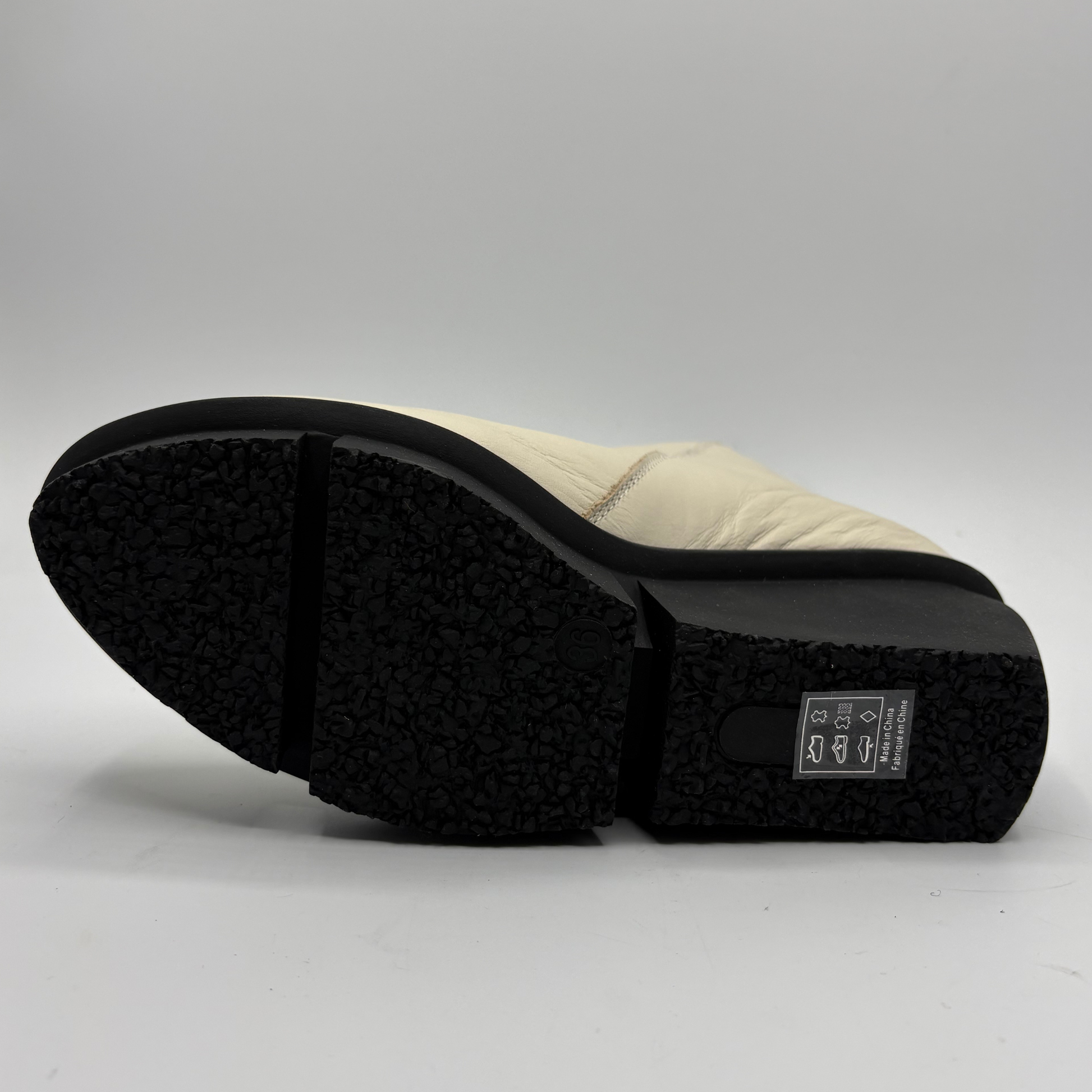 A close-up shot of the sole of a light beige boot, showing a black textured rubber outsole and a small black brand label on the heel.