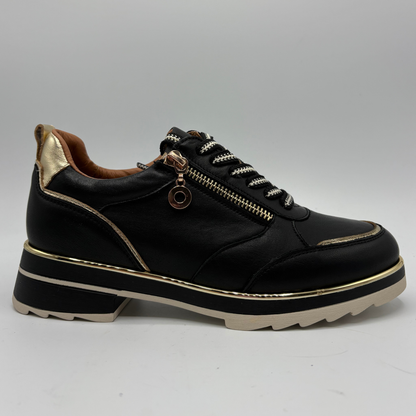 A black leather low-top sneaker with a gold metallic heel tab, gold zipper detail, and gold piping accents, featuring a chunky black and beige platform sole.
