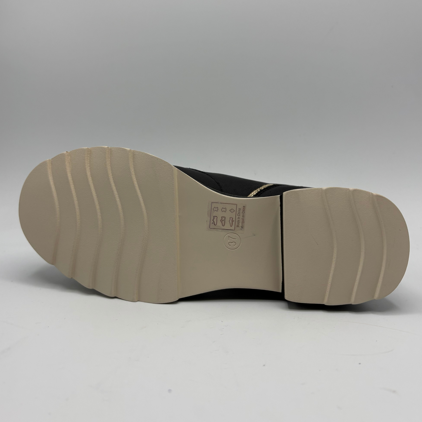 The bottom sole of a black boot or shoe, showing a beige rubber outsole with wavy tread patterns and a small printed size/information label near the center, photographed against a neutral gray studio background.