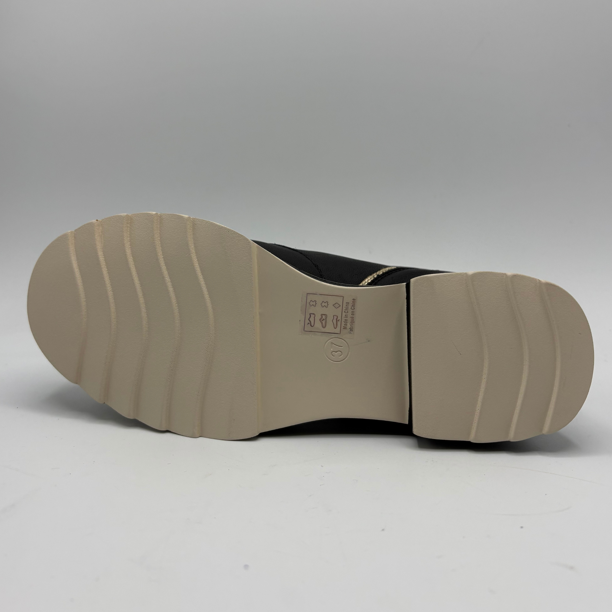 The bottom sole of a black boot or shoe, showing a beige rubber outsole with wavy tread patterns and a small printed size/information label near the center, photographed against a neutral gray studio background.
