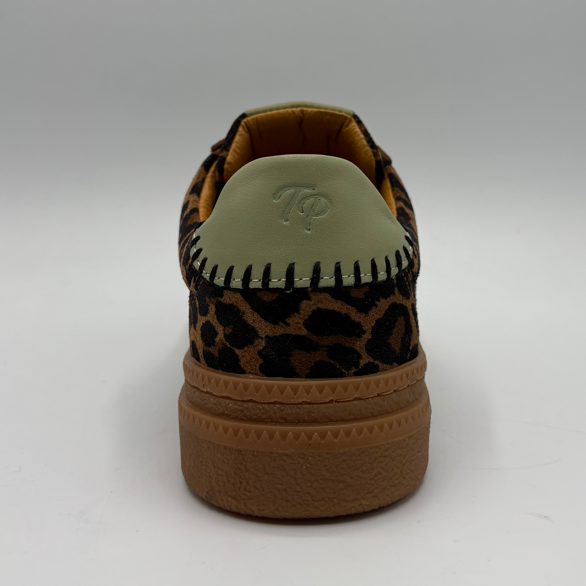 A single low-top sneaker shown from the back on a light gray studio background. The shoe features a brown and black leopard print upper, a light olive green heel tab with an embossed 'TP' logo, and a thick brown gum sole.