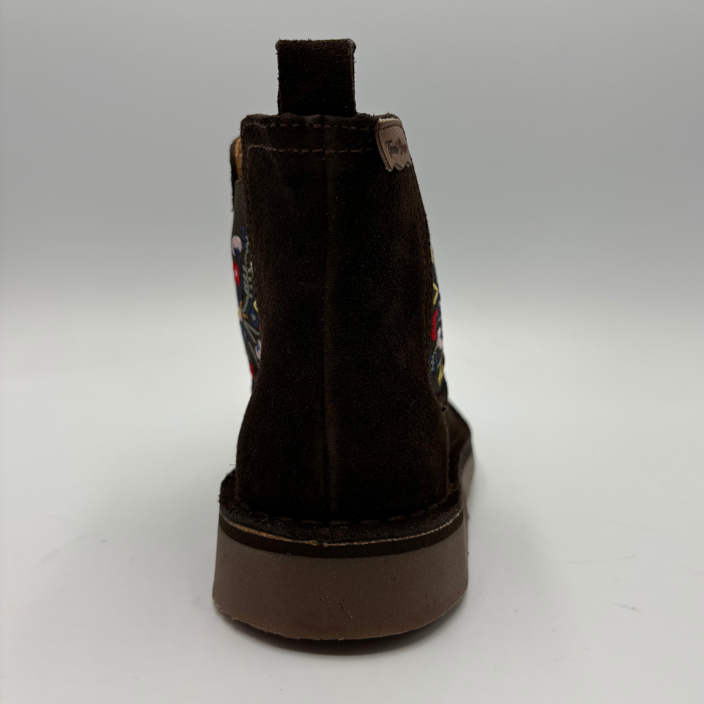 A dark brown suede ankle boot shown from the back, featuring colorful embroidered details on the side and a thick brown sole, set against a neutral gray background.