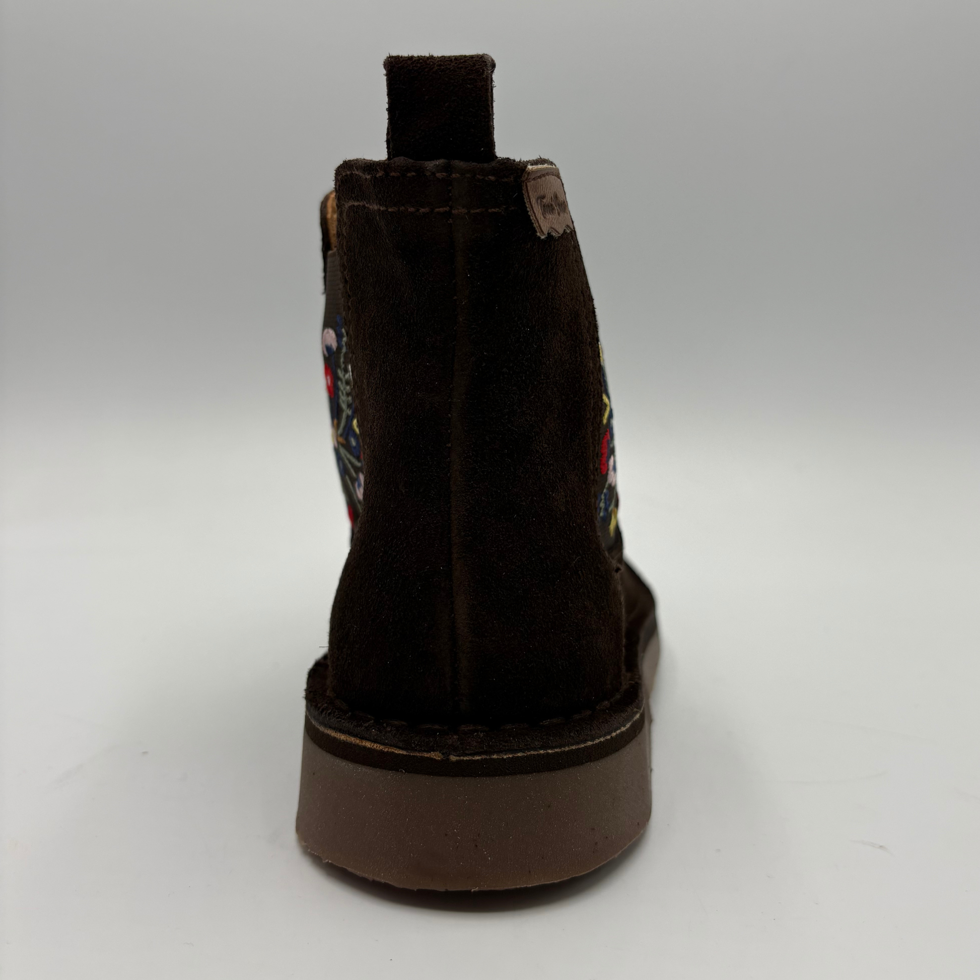 A dark brown suede ankle boot shown from the back, featuring colorful embroidered details on the side and a thick brown sole, set against a neutral gray background.