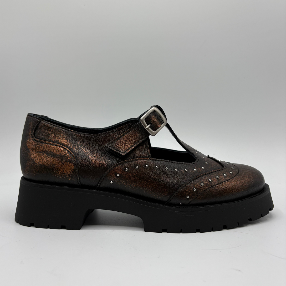 A single brown distressed leather T-strap Mary Jane shoe with silver stud detailing and a black chunky lug sole, shown from a side profile on a light gray background.