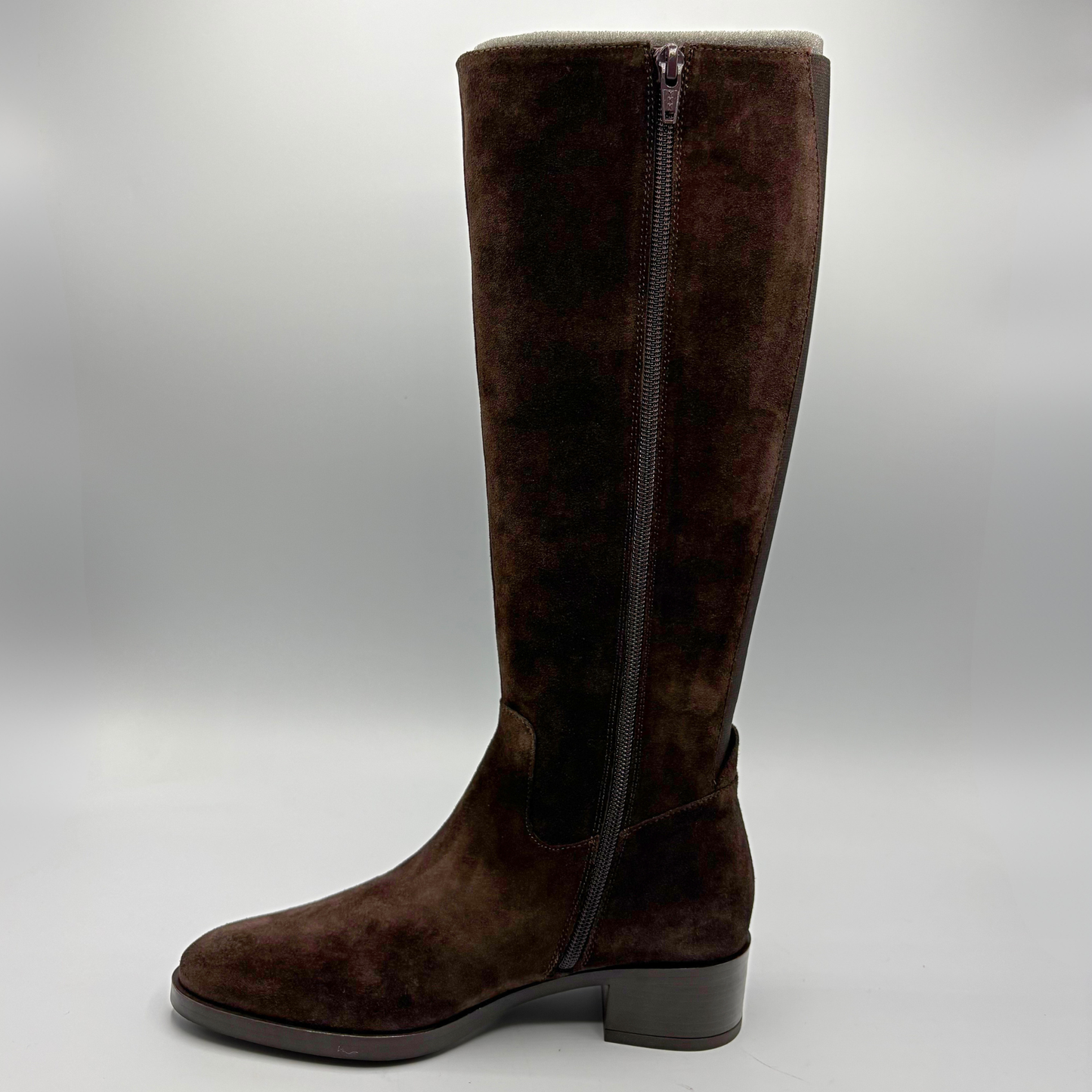 A dark brown suede knee-high boot with a side zipper, low block heel, and rounded toe, shown against a neutral gray background.