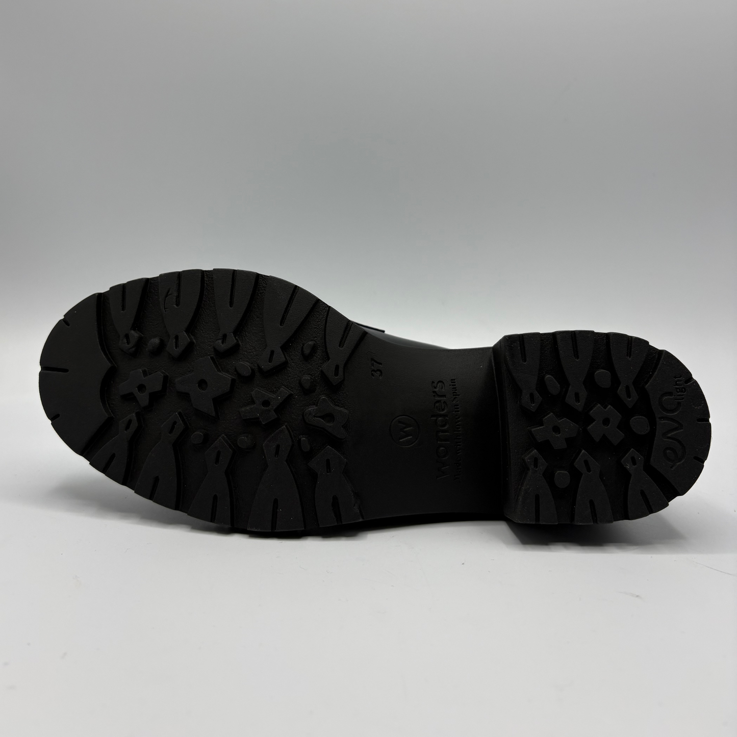 The black rubber outsole of a shoe, showing deep tread patterns and embossed branding, photographed against a neutral gray background.