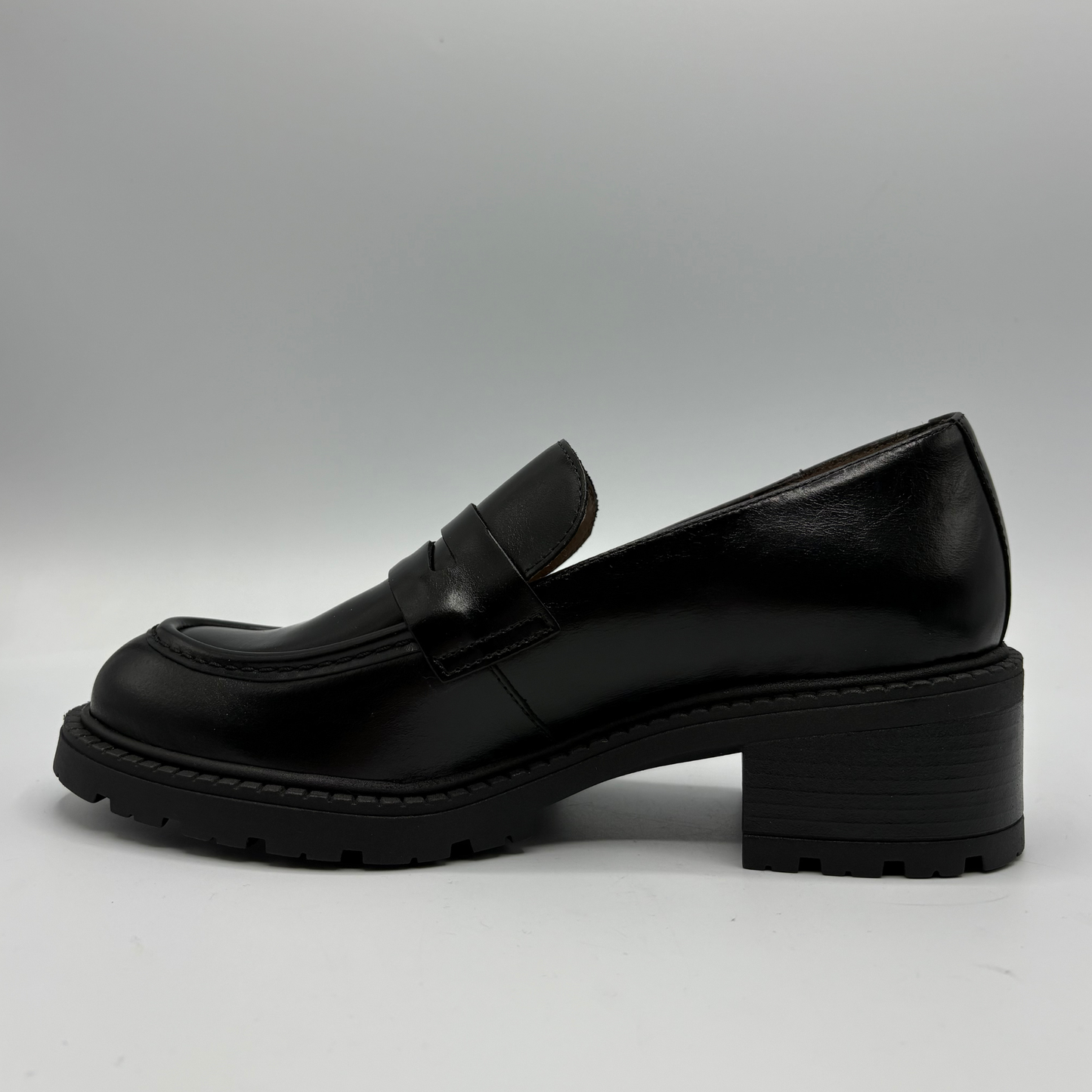 A single black leather penny loafer with a chunky block heel and lug sole, shown in a side profile against a neutral gray background.
