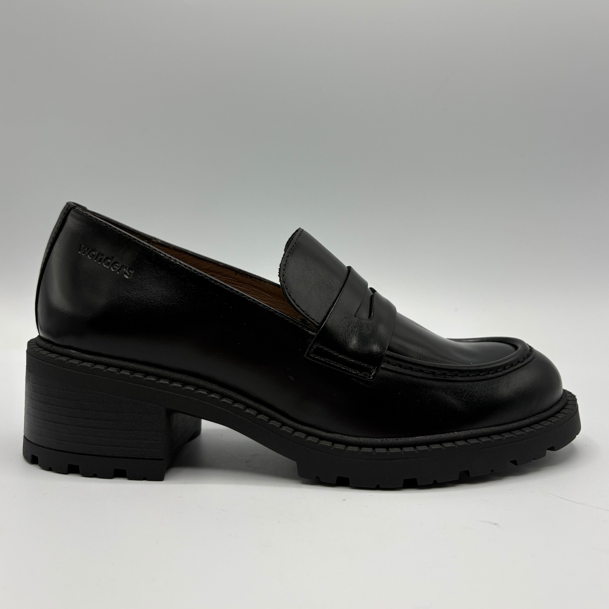 A single black leather penny loafer with a chunky block heel and lug sole, shown in profile against a neutral gray background.